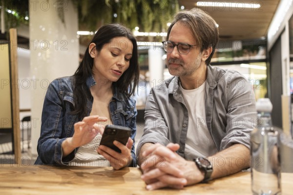 A couple sits at a wooden table, engaged in a lively conversation while sharing something on a smartphone. They appear relaxed and happy in a warmly lit indoor setting