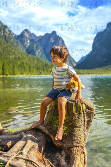 Young boy sitting on a tree stump by lake dobbiaco, holding his plush toy and admiring the dolomites landscape