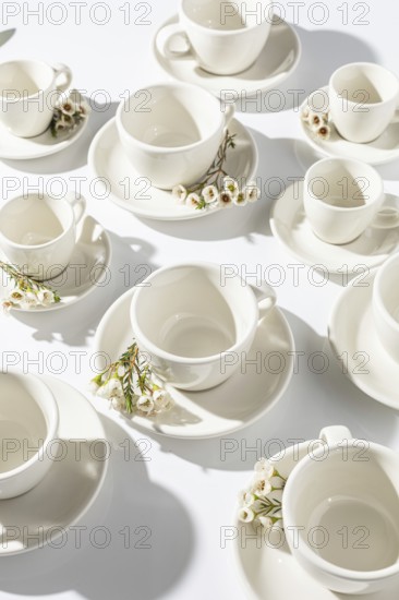 Top view of a tasteful arrangement of white coffee cups and saucers, adorned with delicate spring white flowers, presented on a bright background for a fresh, aesthetic look