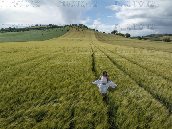 A young woman strolls through lush green fields under a wide blue sky in rural Italy, embracing the natural beauty and tranquility of the picturesque landscape around her