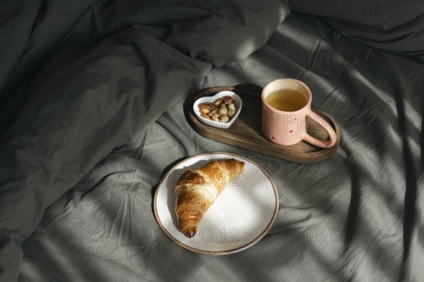 A golden homemade croissant rests on a white plate, accompanied by a wooden tray holding a pink cup of tea and a heart shaped bowl of nuts, all set on a cozy grey bed