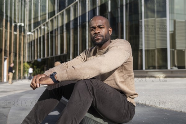 A cuban man in casual attire sits outside a modern glass building, embodying a relaxed urban vibe. Sunlight highlights the architectural lines and his poised demeanor