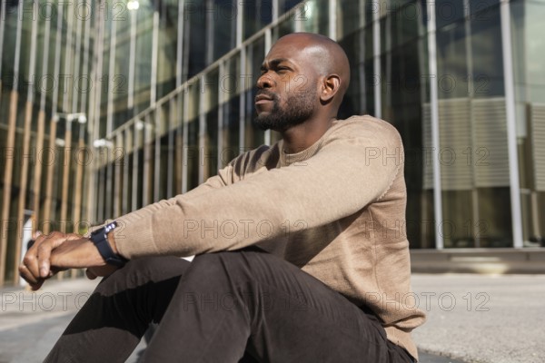 A cuban man in a casual outfit sits outdoors, leaning against a modern glass building. He appears thoughtful and relaxed, enjoying the sunny day in an urban setting