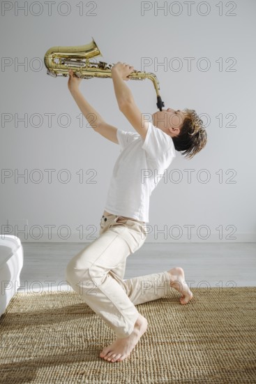 A young boy kneels inside a bright room, passionately playing a saxophone. The scene captures the joyful energy and enthusiasm of musical performance