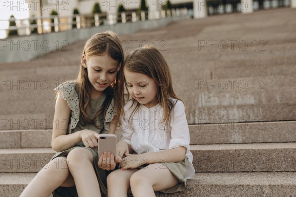 Two young girls sit together on outdoor steps in summer, engrossed in a smartphone. They share a moment of curiosity and exploration, embodying friendship and technology