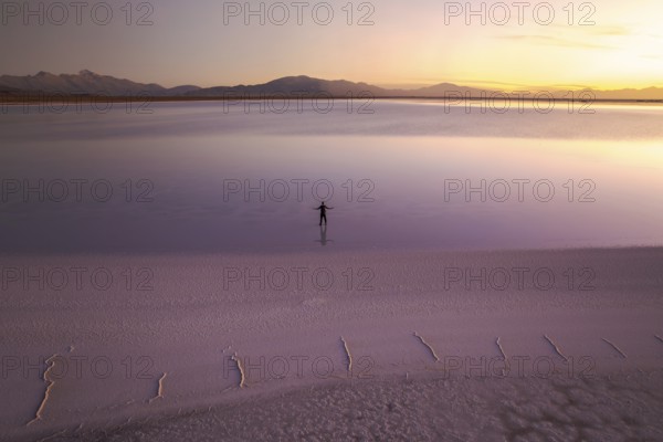A solitary figure stands in the vast expanse of Salinas Grandes, Salta, Argentina, at sunset. The peaceful scene features a pastel sky reflected on the surface of the salt flat