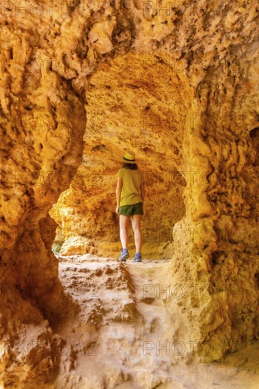A woman stands in a cave with a yellow rock wall. She is wearing a green hat and shorts
