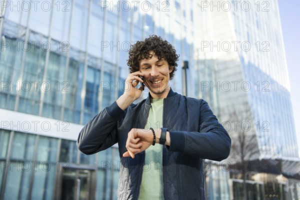 Smiling man in casual attire checks the time on his smartwatch while talking on a smartphone. Modern glass buildings form the urban cityscape backdrop, symbolizing connectivity and business