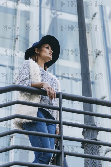 Woman in a stylish hat and fur vest stands confidently on a urban balcony. Blue jeans and a modern glass building in the background emphasize a chic urban look