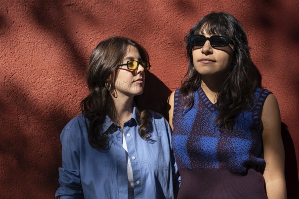 A multiethnic lesbian couple shares a moment outdoors, standing against a sunlit red wall. Both women, in stylish sunglasses, embrace diversity, pride, and love in their picturesque setting