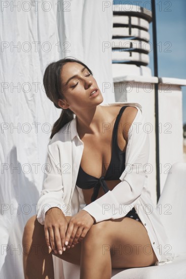 A young woman in stylish swimwear and a white shirt enjoys the summer sun on a rooftop The bright sky and serene atmosphere highlight her relaxed mood