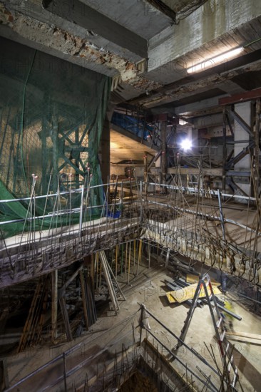 An interior construction site with exposed beams, scaffolding, and safety nets. Illuminated by bright lights, it shows progress and materials in a dynamic work environment