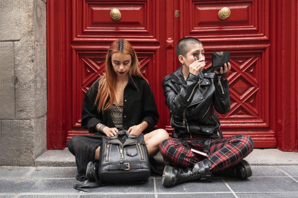 A lesbian couple sits together in front of a red door, each applying makeup with focus and care, highlighting a sense of individuality and shared moments