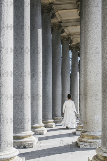 A young girl walks gracefully among towering columns, dressed for her First Communion. The sunlight enhances the serene atmosphere and highlights the elegance of the moment