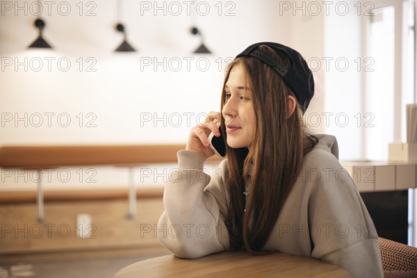 A teenage girl in a cozy coffee shop is engaged with her smartphone, wearing a casual hoodie and a backward cap, enjoying a moment of relaxation and connectivity