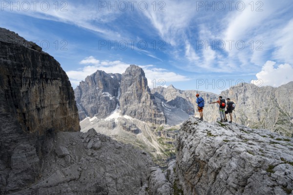 Three mountaineers on a rock in front of a picturesque mountain landscape with rocky peaks, Via Ferrata SOSAT via ferrata, summit of Cima Tosa in the back, Brenta Mountains, Trentino, Italy