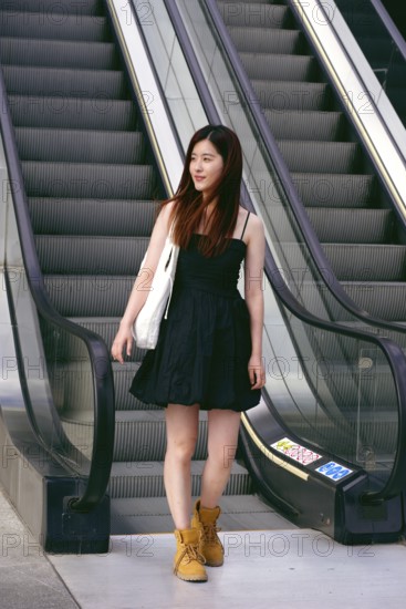 A young asian woman in a black dress stands confidently on an escalator. Her brown hair is loose, and she carries a white bag, highlighting a casual yet stylish urban look