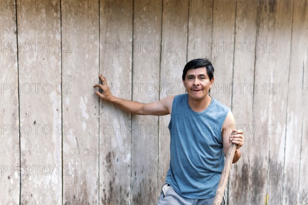 A middle-aged man stands confidently in the Colombian countryside, leaning against a rustic wooden wall while taking a break from miscellaneous farm work