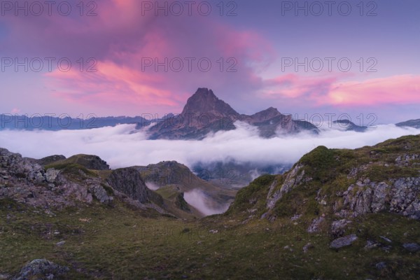 A breathtaking mountain peak surrounded by clouds at sunrise in the Pyrenees. The vibrant pink sky adds a dramatic contrast, reflecting on the serene landscape below in summer