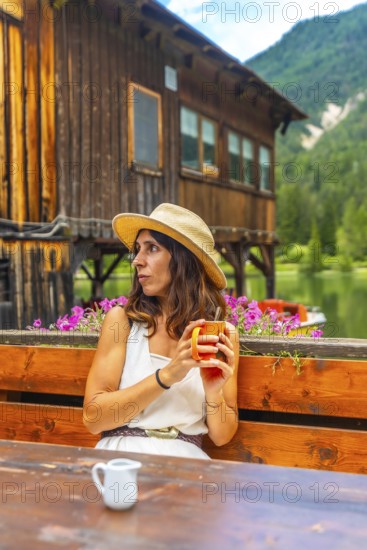 Young woman wearing a straw hat enjoying a hot drink in a mug at a wooden table with a view of lake dobbiaco and the dolomites