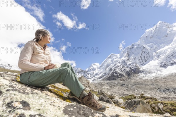 A woman sitting on a rock during a trek in the Everest region, admiring the majestic snow-capped mountains under a clear blue sky. Perfect scene of adventure and serenity