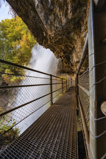 A narrow metal footbridge leads past a large waterfall, flanked by rocks and autumn vegetation, Lake Brienz, Giessbach Waterfall, Switzerland