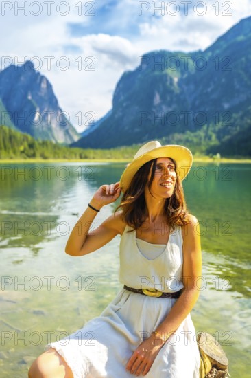Young woman wearing a straw hat relaxing at lake dobbiaco with the dolomites in the background, enjoying summer holidays in italy