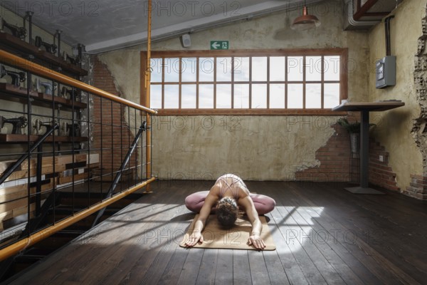 A woman practices yoga on a mat in a rustic loft with wooden floors, surrounded by vintage sewing machines. Sunlight streams through large windows, casting gentle shadows