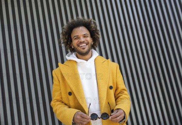 A cheerful Afro man in a yellow coat holds sunglasses, standing in front of a modern striped backdrop. His confident smile and casual attire highlight contemporary urban style