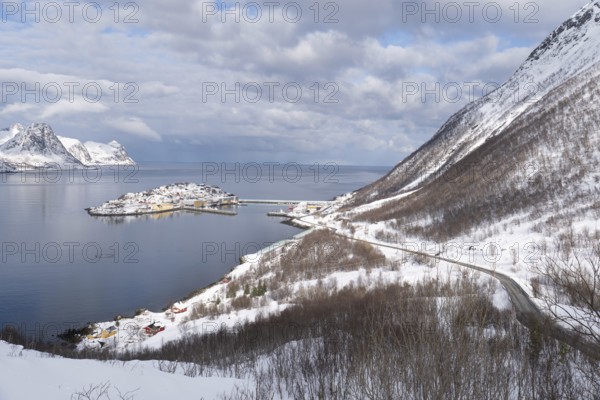 Serene winter view of Husoy, Norway, with a snow-covered coastline, calm sea, and dramatic mountains under a partly cloudy sky. A picturesque northern village in white