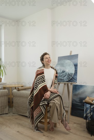 A woman sits in a bright, cozy home art studio with a colorful blanket draped over her shoulders, smiling. Paintings are displayed on easels, creating a warm atmosphere