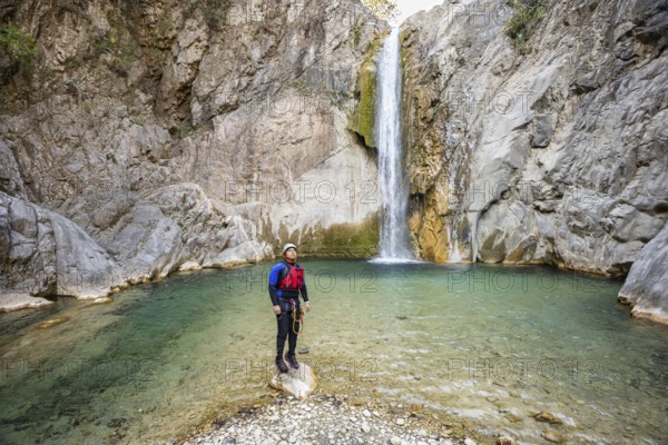 Adventurer stands on a rock in the middle of a clear stream facing a tall waterfall in Matacanes, Nuevo Leon, Mexico. Surrounded by rugged canyon cliffs and nature