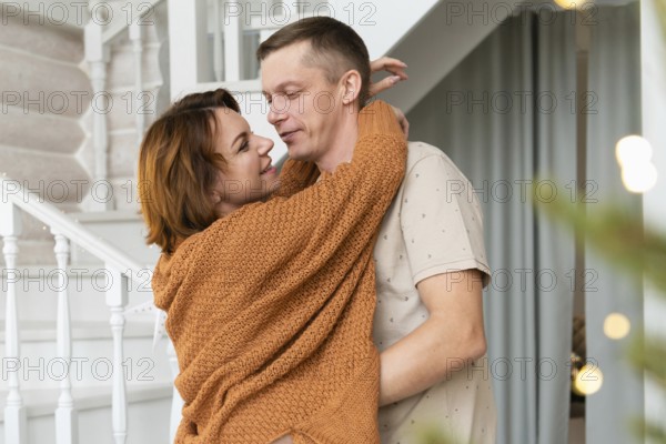 A warm embrace between a man and a woman celebrating Christmas. Both are smiling gently, facing each other. The setting features a festive background with a hint of holiday decorations and a staircase