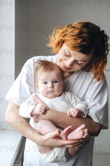A mother lovingly holds her newborn baby girl, captured in warm natural light. The white clothing enhances the gentle and tender moment shared between them