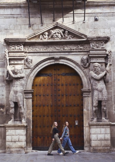 Two young woman friends casually walk past an ornate wooden door framed by intricate stone carvings. The architectural details highlight a blend of history and modernity