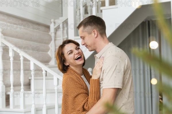 A joyful woman and a smiling man are engaging closely in a warm embrace in an elegantly decorated home, sharing a moment of laughter by a staircase
