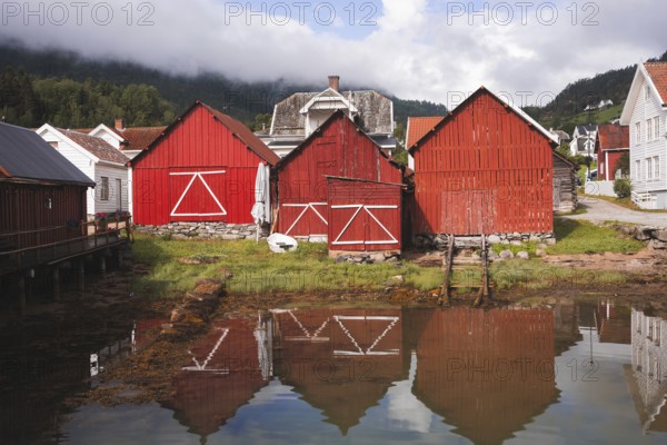 Charming red wooden huts reflect in the calm water of a Norwegian fjord village Surrounded by lush greenery and misty mountains, perfect for outdoor adventures, trekking, or camping