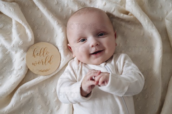 A smiling baby boy lies on a soft, cream blanket. Beside him, a wooden plaque reads Hello World, capturing the warmth and innocence of early childhood moments