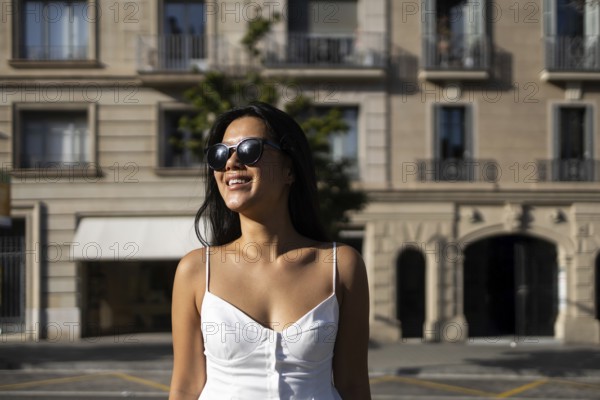 A fashionable Chinese woman in sunglasses and a white dress enjoys a bright summer day in the city, standing against a backdrop of urban architecture and basking in the sunlight