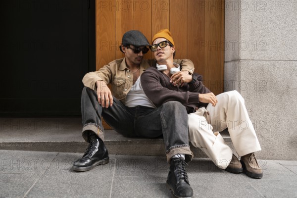 A Colombian gay couple sits on a city street, embracing while enjoying a casual moment. Wearing stylish clothing and sunglasses, they exude happiness and connection in an urban setting