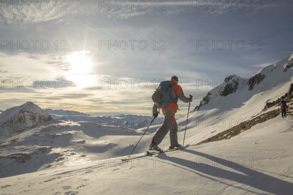 Two backcountry skiers ski down a pristine snowy mountain under a bright sky. Equipped with backpacks and poles, they master the slope with determination and skill