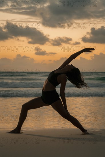 A woman gracefully poses in a yoga stance on a beach during sunset in Tulum, Mexico. The serene ocean waves and vivid sky create a tranquil backdrop for her meditative practice
