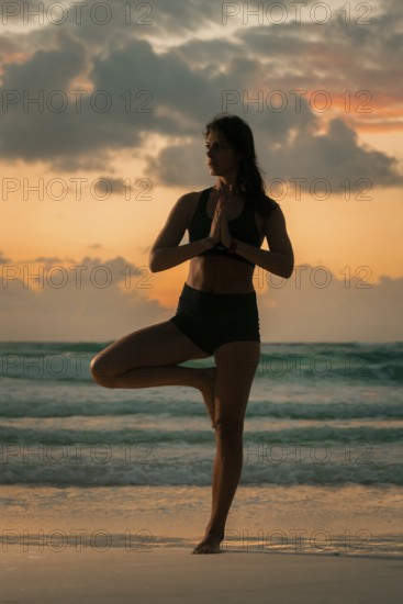 A woman performs a yoga pose on a tranquil beach as the sun sets, blending calm and focus against a colorful sky, symbolizing peace and strength in nature of Tulum, Mexico