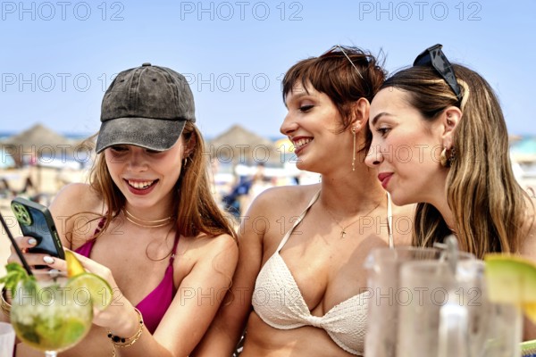 Three friends relax at a sunny beach, sipping colorful cocktails and checking their phones. The warm weather creates an inviting summer vibe with laughter and joy all around