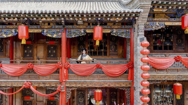 A woman dressed in traditional Qing dynasty clothing stands on a intricately decorated balcony in Pingyao, China, looking at the camera amidst red lanterns and festive decorations