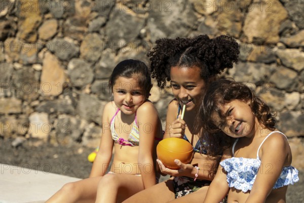 Three children in swimsuits sit happily against a stone wall. They enjoy refreshing drinks, basking in the sunlight, and embodying the joy of summer vacations