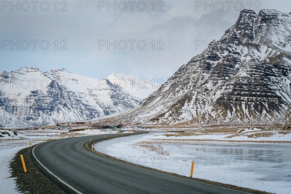 A curved road leads through stunning snowy mountains in Iceland. The winter landscape features rugged peaks under a cloudy sky, creating a serene atmosphere