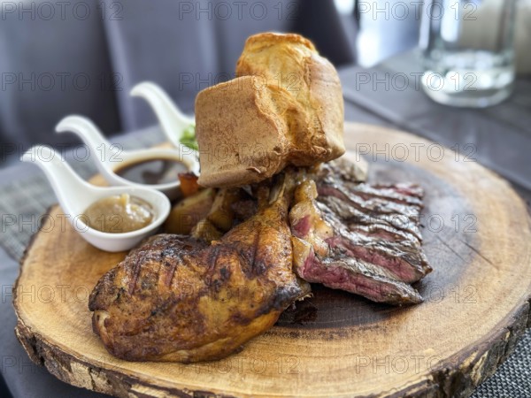 A succulent plate of steak slices and roasted chicken leg served with Yorkshire pudding and a trio of delicious sauces, presented on a rustic wooden board on a cafe in Thailand