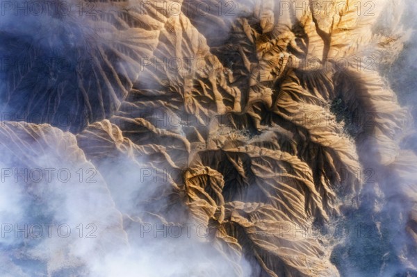 An aerial view showcases rocky mountain formations with deep ridges and valleys Wisps of mist partially obscure the rugged terrain, creating a mystical atmosphere