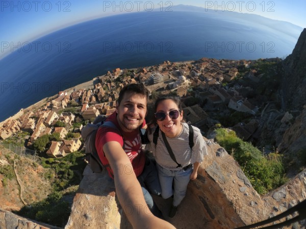 A joyful couple takes a selfie overlooking the charming town and azure sea from the heights of Monemvasia, Greece, on a sunny day. The vibrant landscape offers a perfect backdrop